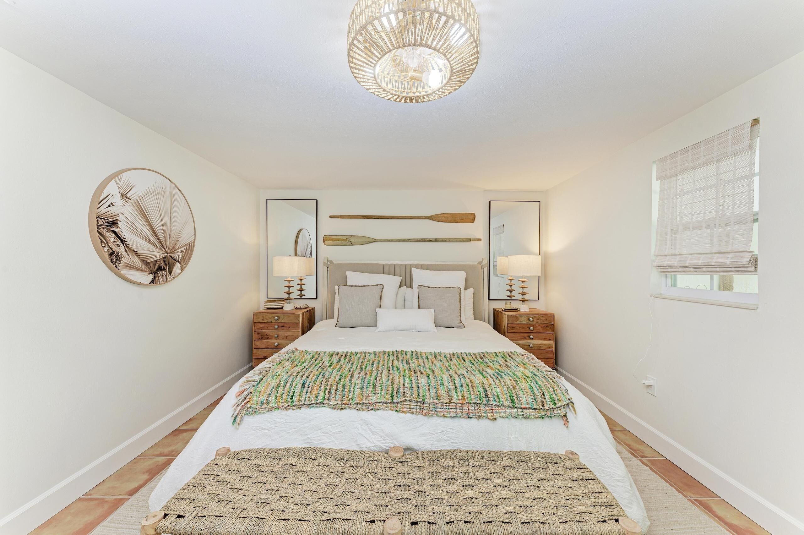 A bedroom with a bed, a large circular wall decoration, two nightstands with lamps, a wooden headboard, and a window with blinds.