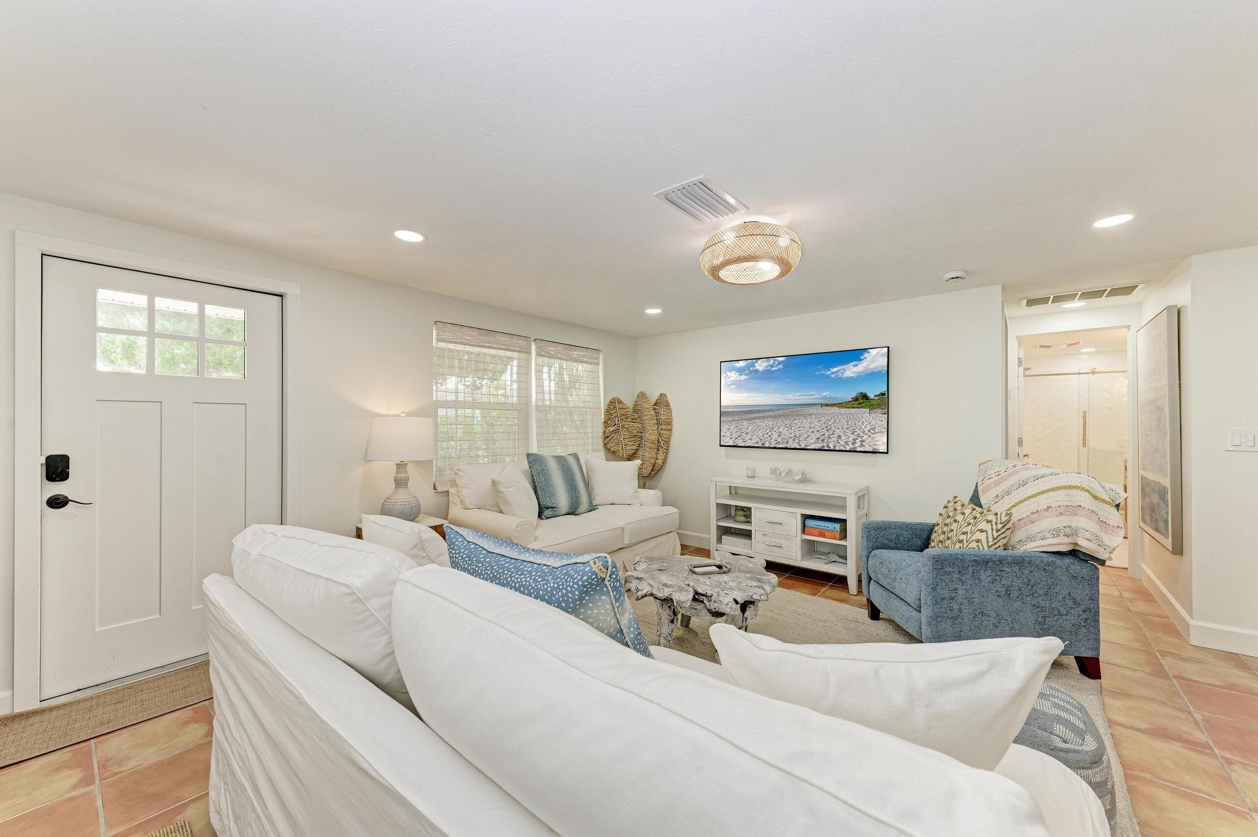 A modern living room with a white sofa, a blue armchair, and a coffee table. There is a large window with blinds, a painting on the wall, and a door with a handle.