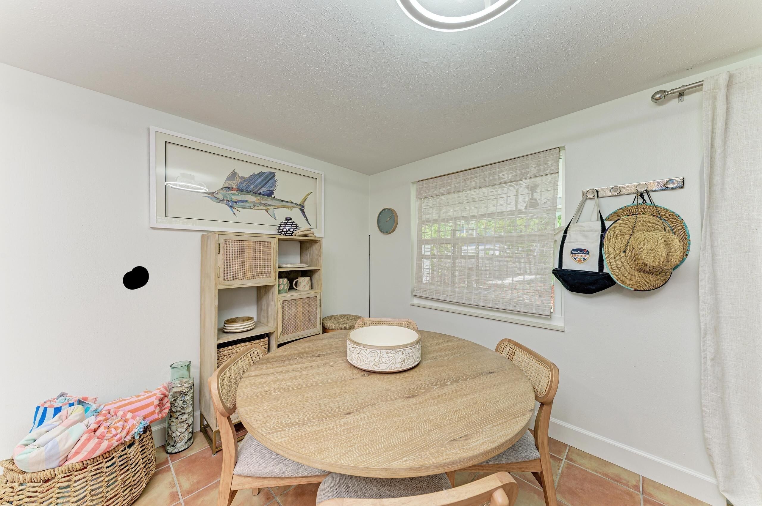 A round wooden table with chairs, a framed artwork on the wall, and a window with curtains.