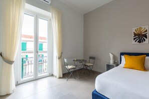 A hotel room with a bed, a small table with chairs, a nightstand, and a view of a balcony with green shutters. - Duomo Charme Studio by Wonderful Italy (Naples)