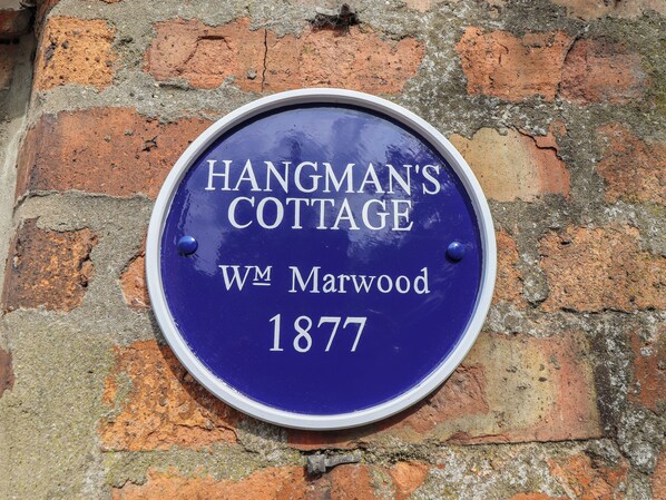 A circular sign with 'HANGMAN'S COTTAGE WM Marwood 1877' written on it, mounted on a brick wall.