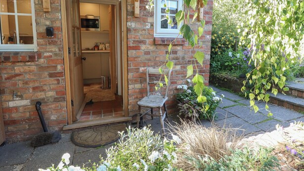 A brick house with a wooden door, a small outdoor seating area, and a garden with various plants. - Hangmans Cottage (Horncastle)