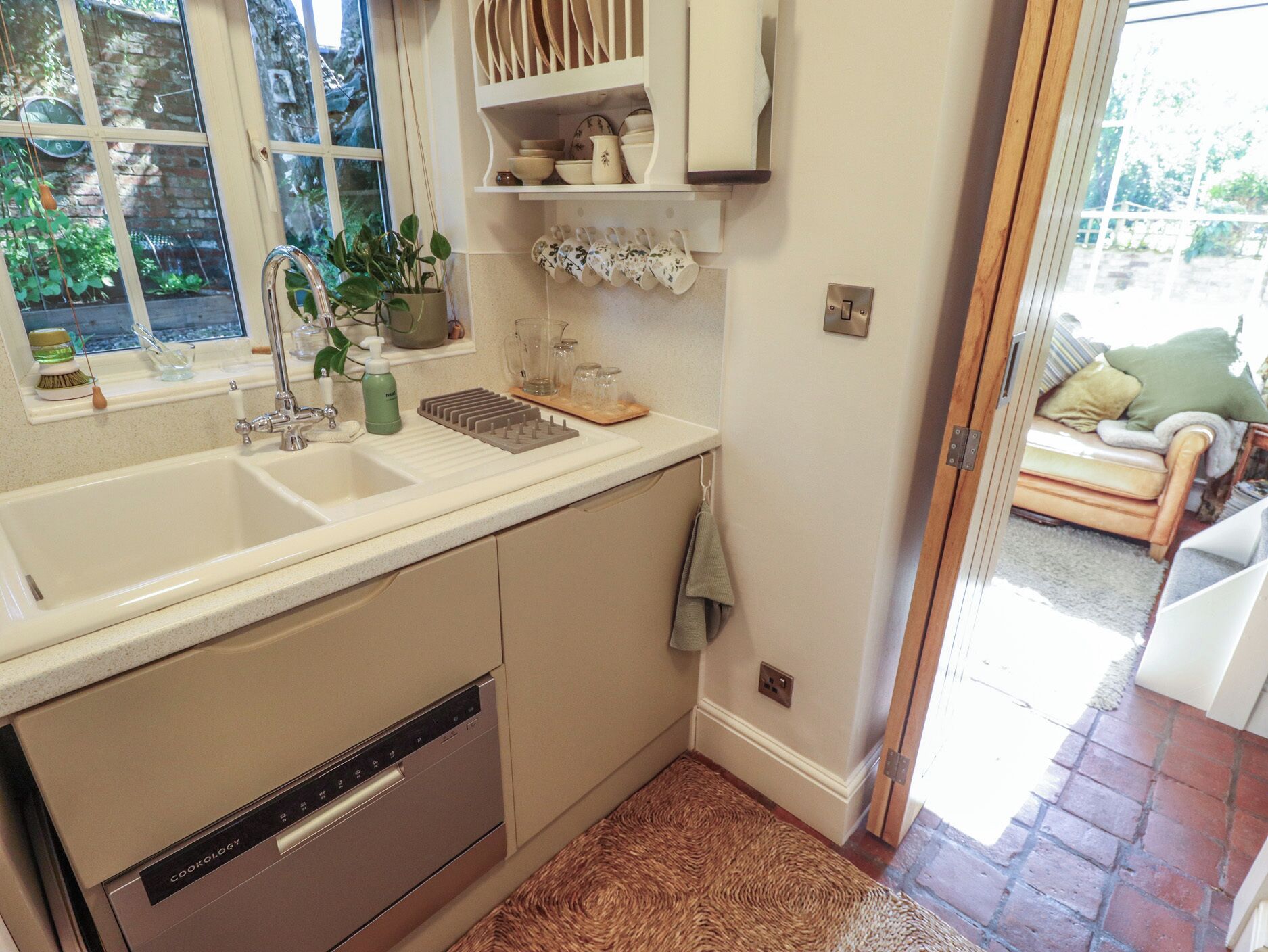 Una cocina con lavavajillas incorporado, una ventana con vista a la vegetación y una puerta que da a una sala de estar.