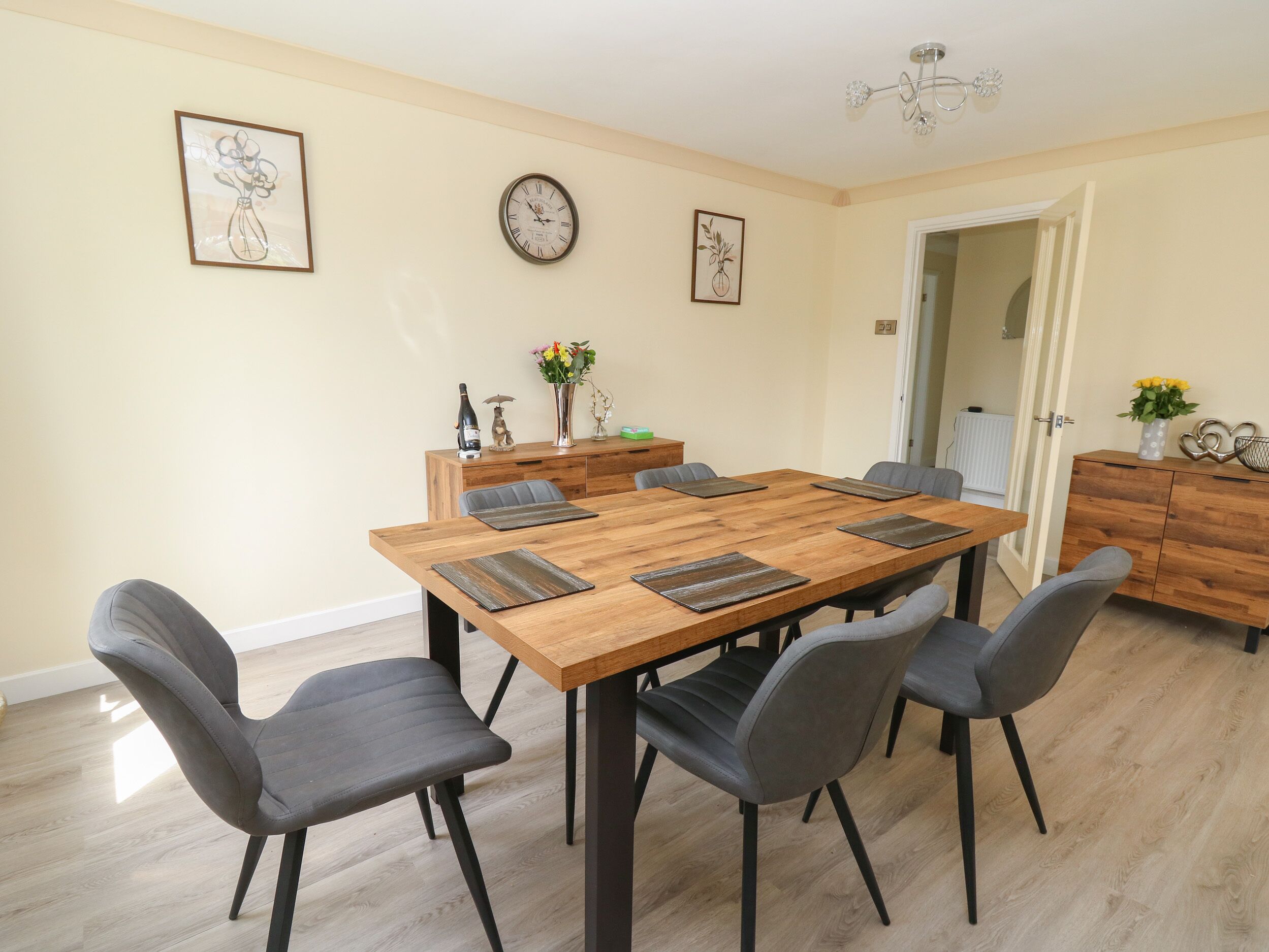 Een eetruimte met een houten tafel, zes grijze stoelen, een houten kast en twee wandklokken.