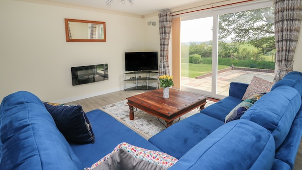 A living room with a large blue sofa set, a flat-screen TV, a wooden coffee table, and a view of a garden through a sliding glass door. - Erwlon (Aberystwyth)