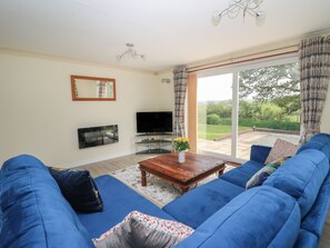 A living room with a large blue sofa set, a flat-screen TV, a wooden coffee table, and a view of a garden through a sliding glass door. - Erwlon (Aberystwyth)