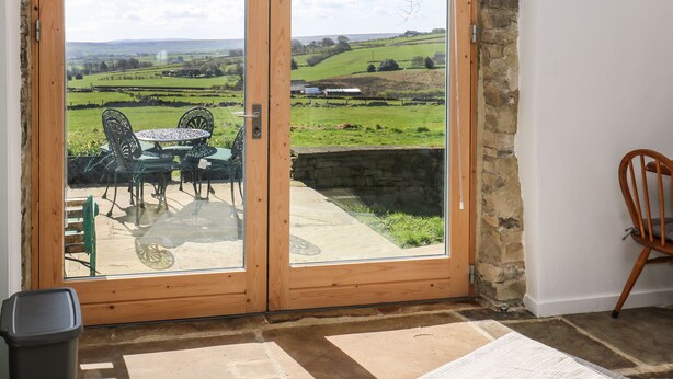 Une porte coulissante à cadre en bois, dotée d’un panneau de verre, qui donne sur un patio extérieur avec une table et des chaises, et offre une vue sur un paysage verdoyant.