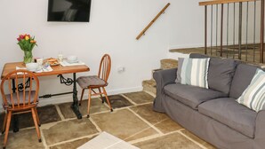 Un salon chaleureux avec un canapé gris, une table à manger en bois et une chaise. Un téléviseur est fixé au mur, et un escalier mène à un autre niveau.
