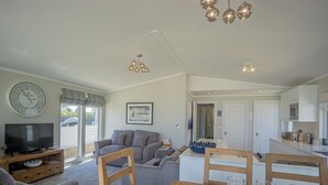 A modern living room with a flat-screen TV, grey sofa, wooden dining table with glass candle holders, and a ceiling light fixture. - E19 The Warren (Pwllheli)