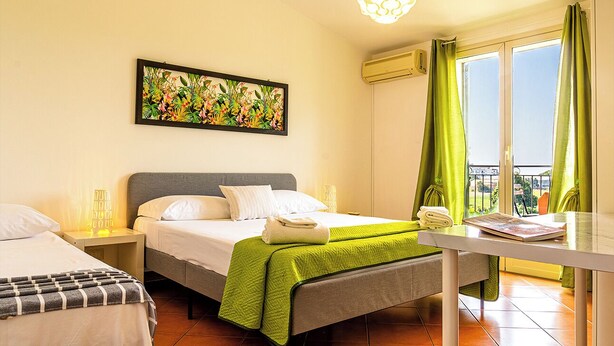 A hotel room with a bed, a bedside table, a lamp, a painting, a window with curtains, and a balcony view. - Villetta Mari e Amuri (Giardini Naxos)