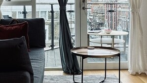 Un salon moderne avec un canapé, une petite table ronde et une vue sur le paysage urbain, visible à travers de grandes fenêtres ornées de rideaux.