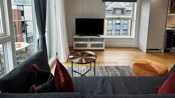 Un salon moderne avec un grand canapé, une table basse et un téléviseur à écran plat. On y trouve une fenêtre avec des rideaux, une vue sur les bâtiments à l’extérieur et un coin cuisine à l’arrière-plan.