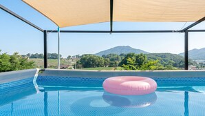 A swimming pool with a pink inflatable ring, surrounded by a shaded area with a view of green hills and mountains. - Ganghwado Labyrinthos Companimal Pension (Incheon)