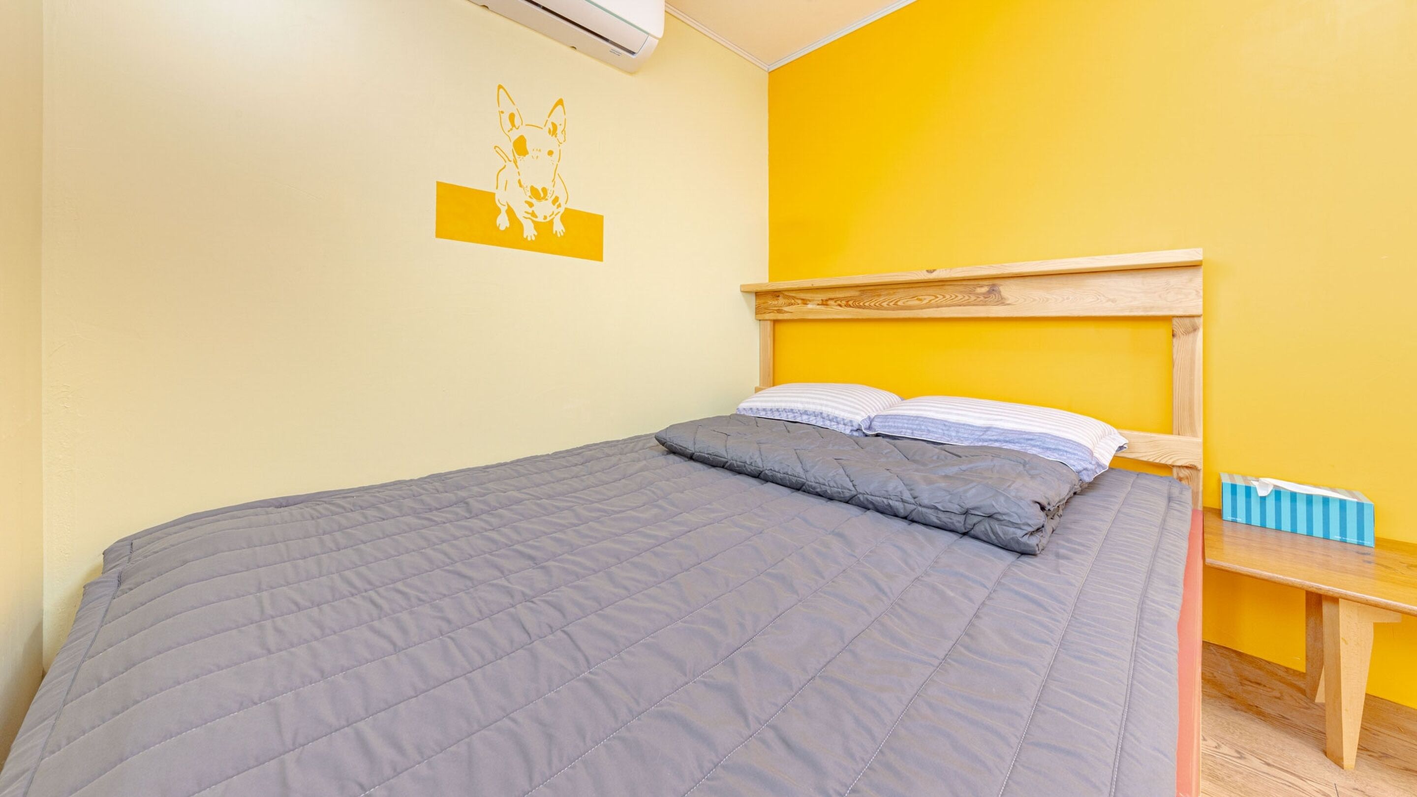 A bed with a grey quilt and pillows, a wooden headboard, a small bedside table with a blue box, and a wall-mounted air conditioner.