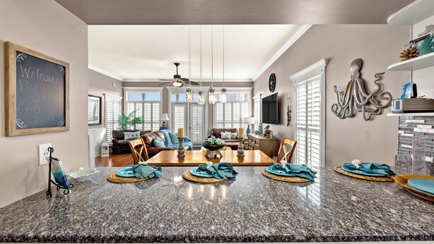 A modern kitchen with granite countertops, a dining area with a wooden table and chairs, and a living room with a sofa and a TV. - Algun Dia - Plush Family-friendly Home - A Block From Beach (Panama City Beach)