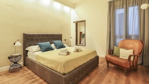 Ein Schlafzimmer mit einem Bett, einem Sessel, einem Nachttisch und einem Fenster mit Vorhängen.