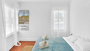 A bedroom with a bed, white curtains, a window with blinds, and a view of greenery outside. - Garden Suite 3-hosted by Sweetstay (Miami Beach)