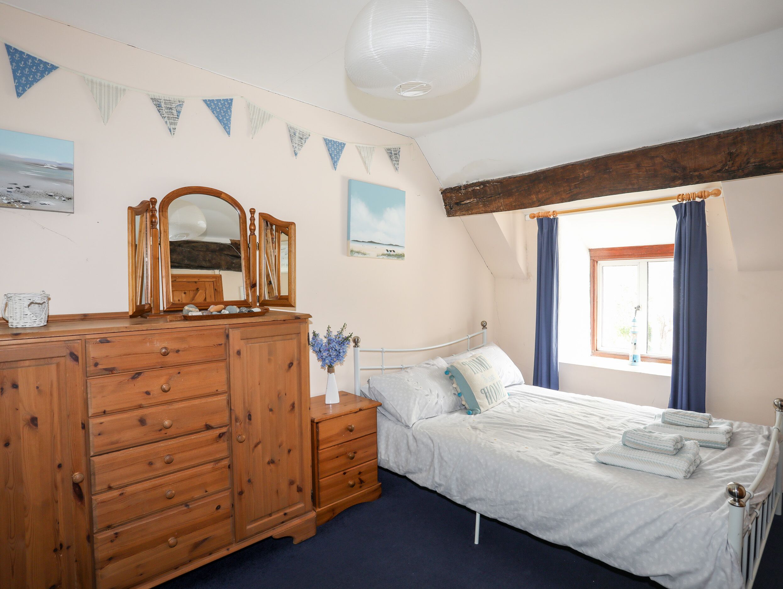 A bedroom with a wooden dresser, a bed with white linens, a mirror, and a window with curtains.