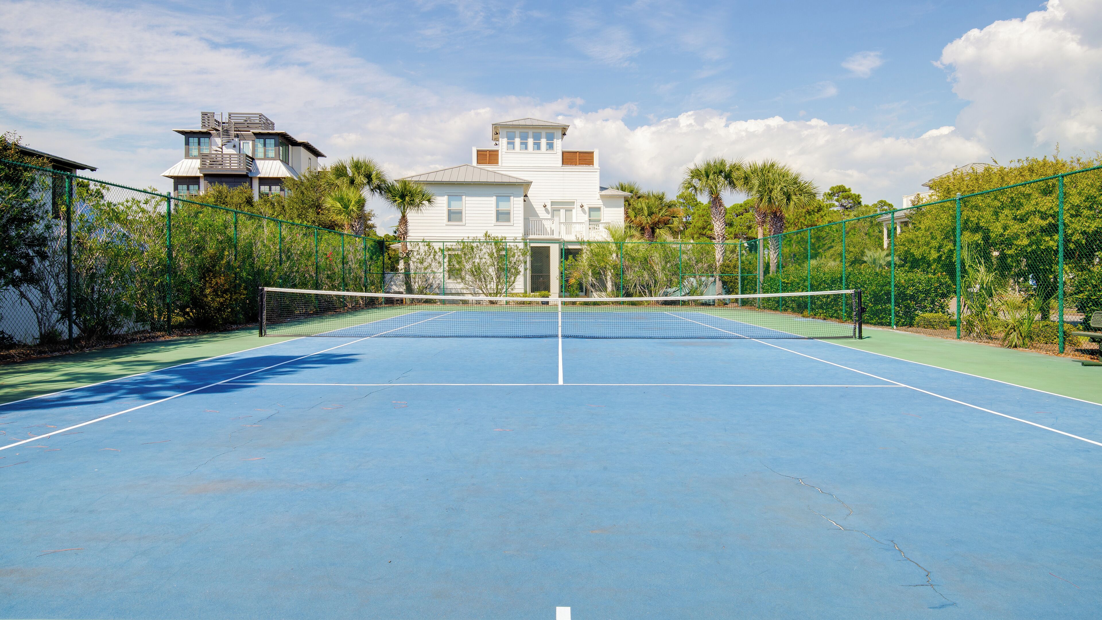 A tennis court with a net, surrounded by green fencing and palm trees.