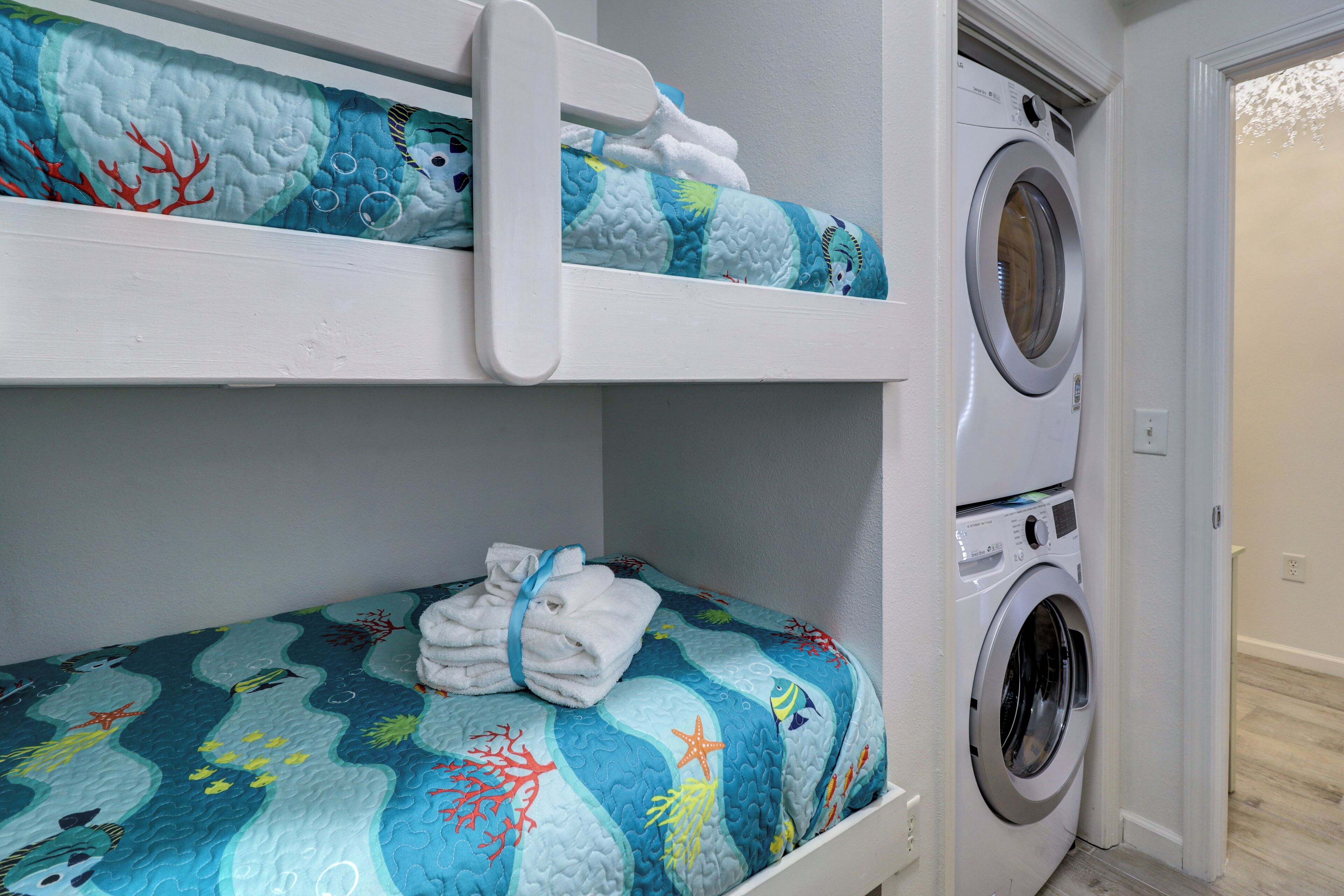 Een stapelbed met blauw-witte beddengoed met een oceaanmotief, een wasmachine en een deur naar een andere kamer.