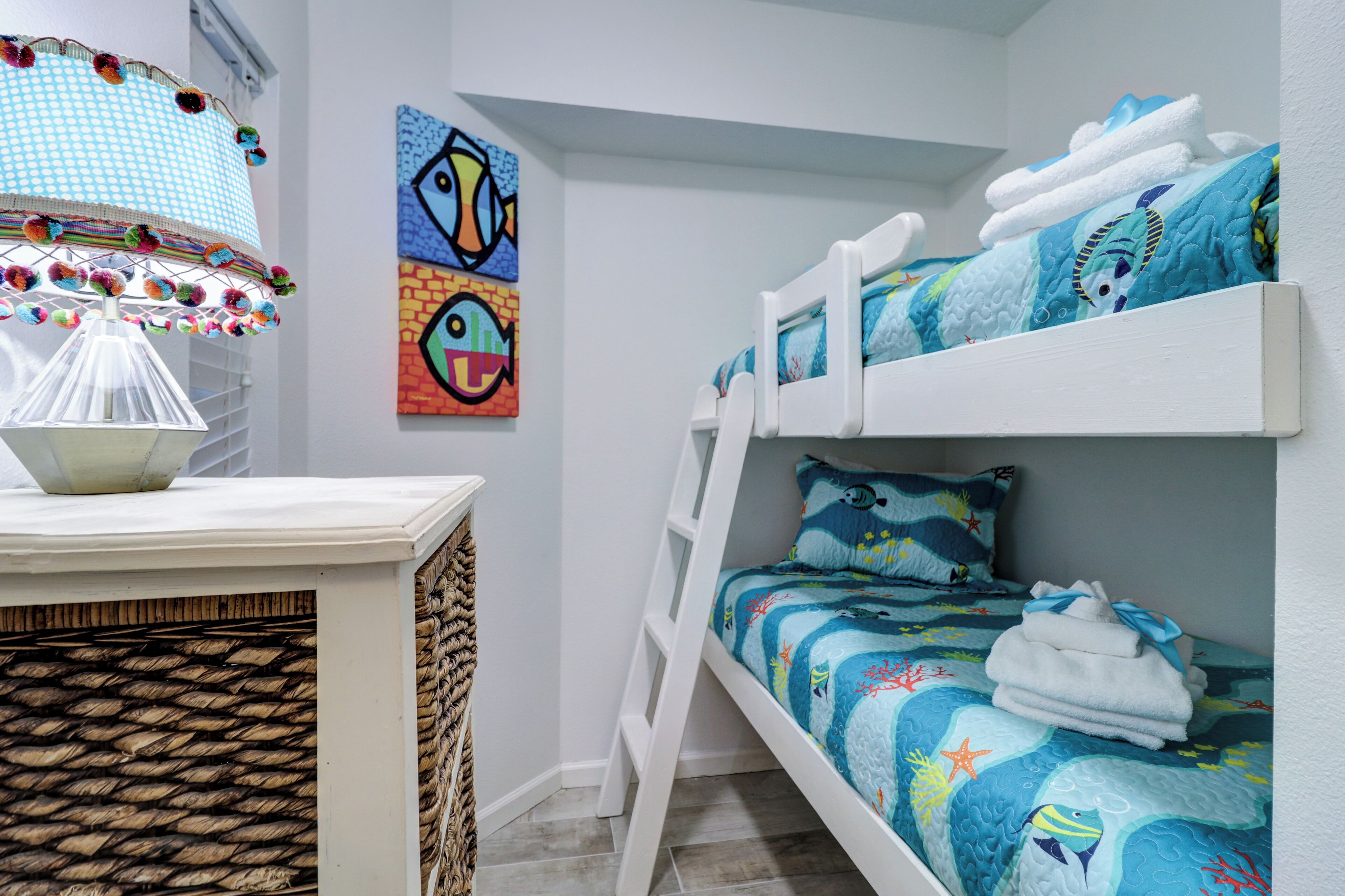 A bunk bed with a blue ocean-themed bedding, a white ladder, and a wall with colorful fish paintings.