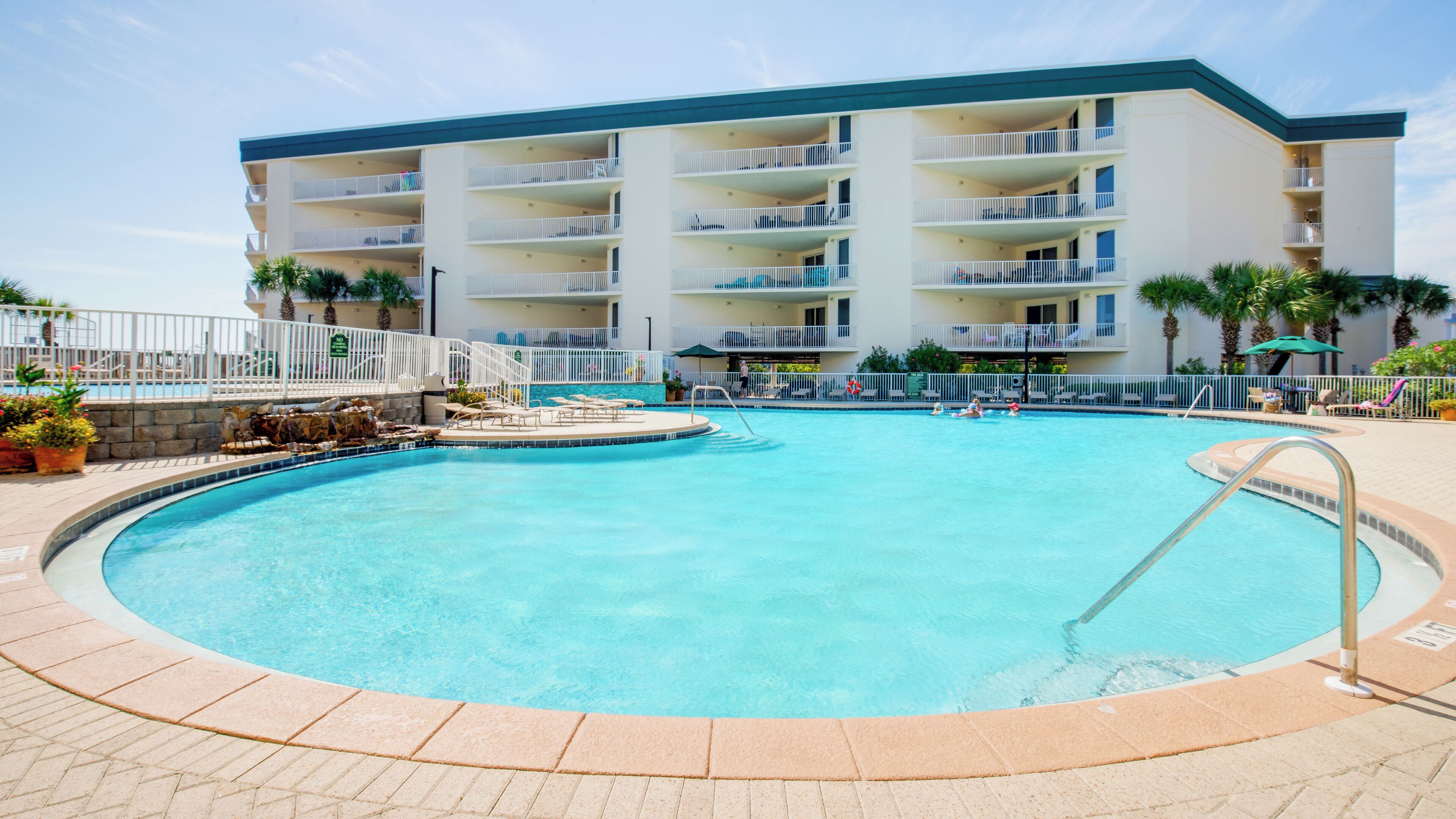 A hotel with a large swimming pool, multiple balconies, and a well-maintained outdoor area with palm trees.