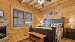 A cozy log cabin bedroom with a stone fireplace, wooden walls, a bed with a plaid bedspread, a bench, and a ceiling fan. - Theatre in the Mountains by Jackson Mountain Rentals (Sevierville)