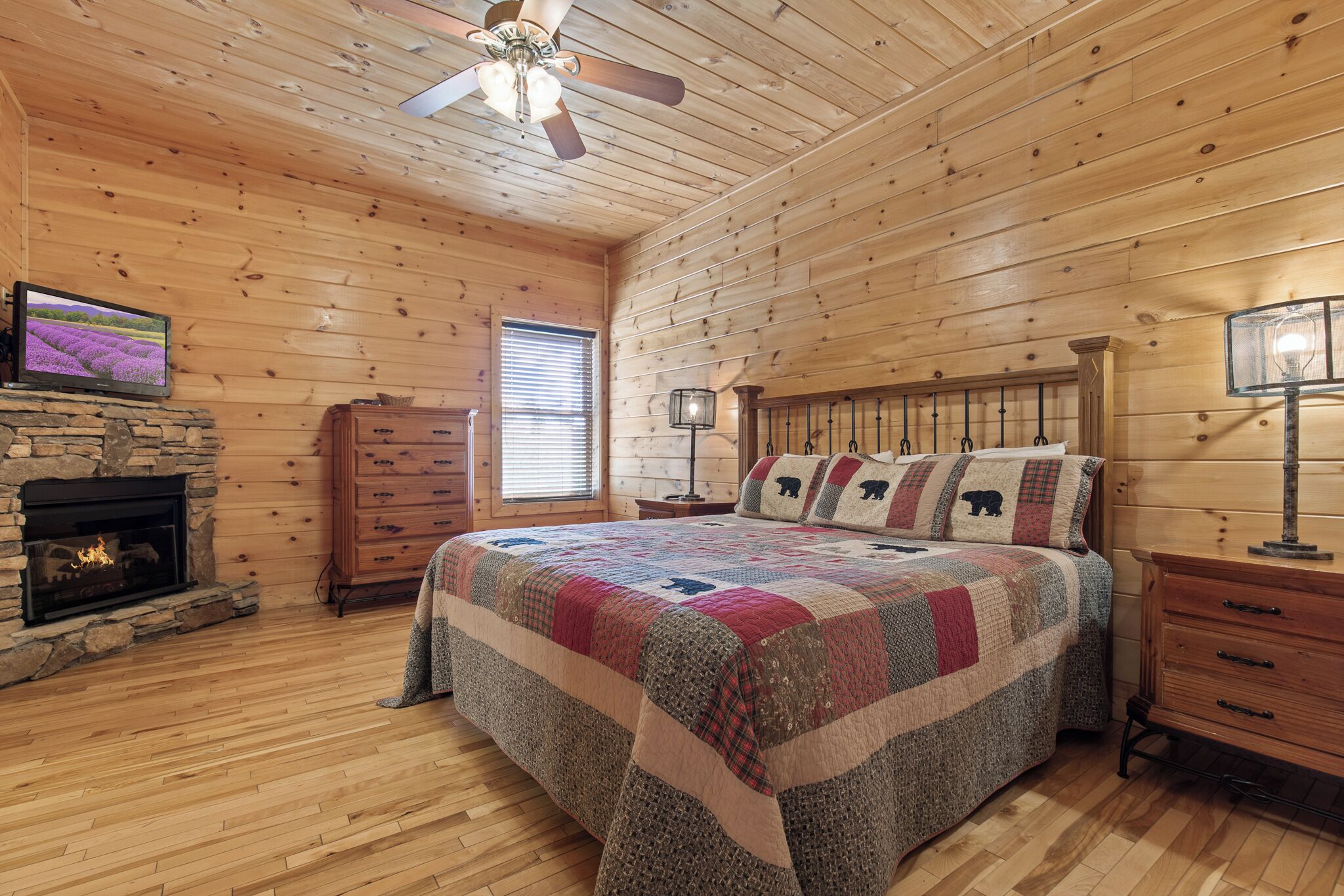 A log cabin bedroom with a stone fireplace, wooden walls, a bed with a quilt, a ceiling fan, and a TV on the wall.