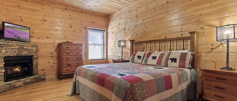 Ein Schlafzimmer in einer Blockhütte mit Kamin aus Stein, Holzwänden, einem Bett mit Quilzudecke, Deckenventilator und Fernseher an der Wand.