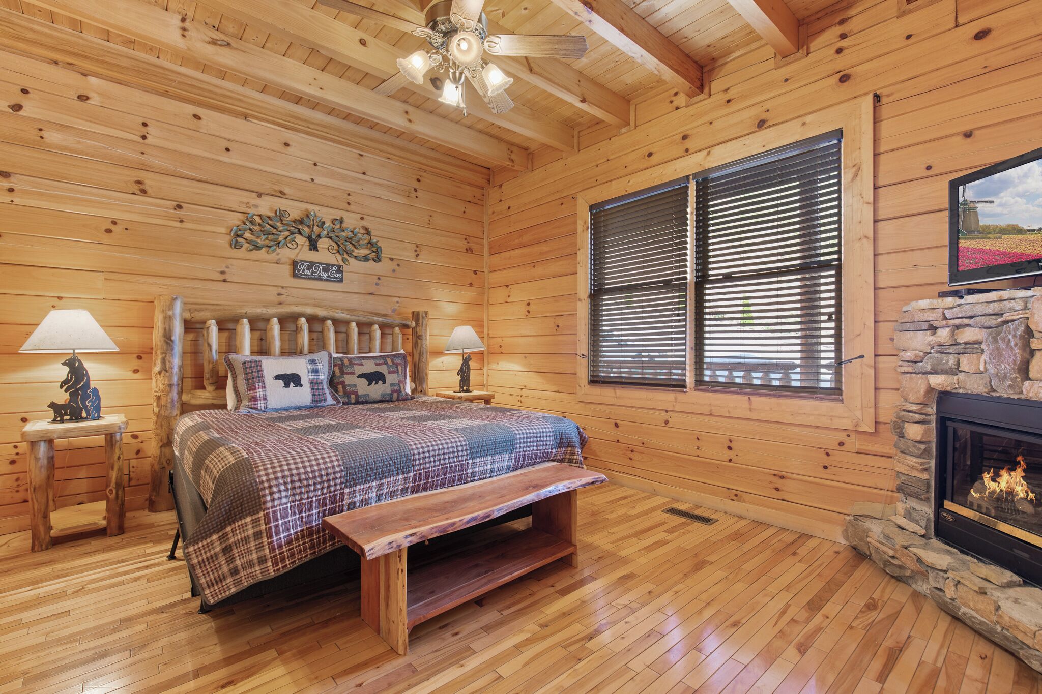 A cozy wooden cabin bedroom with a bed, a stone fireplace, and a large window with blinds.