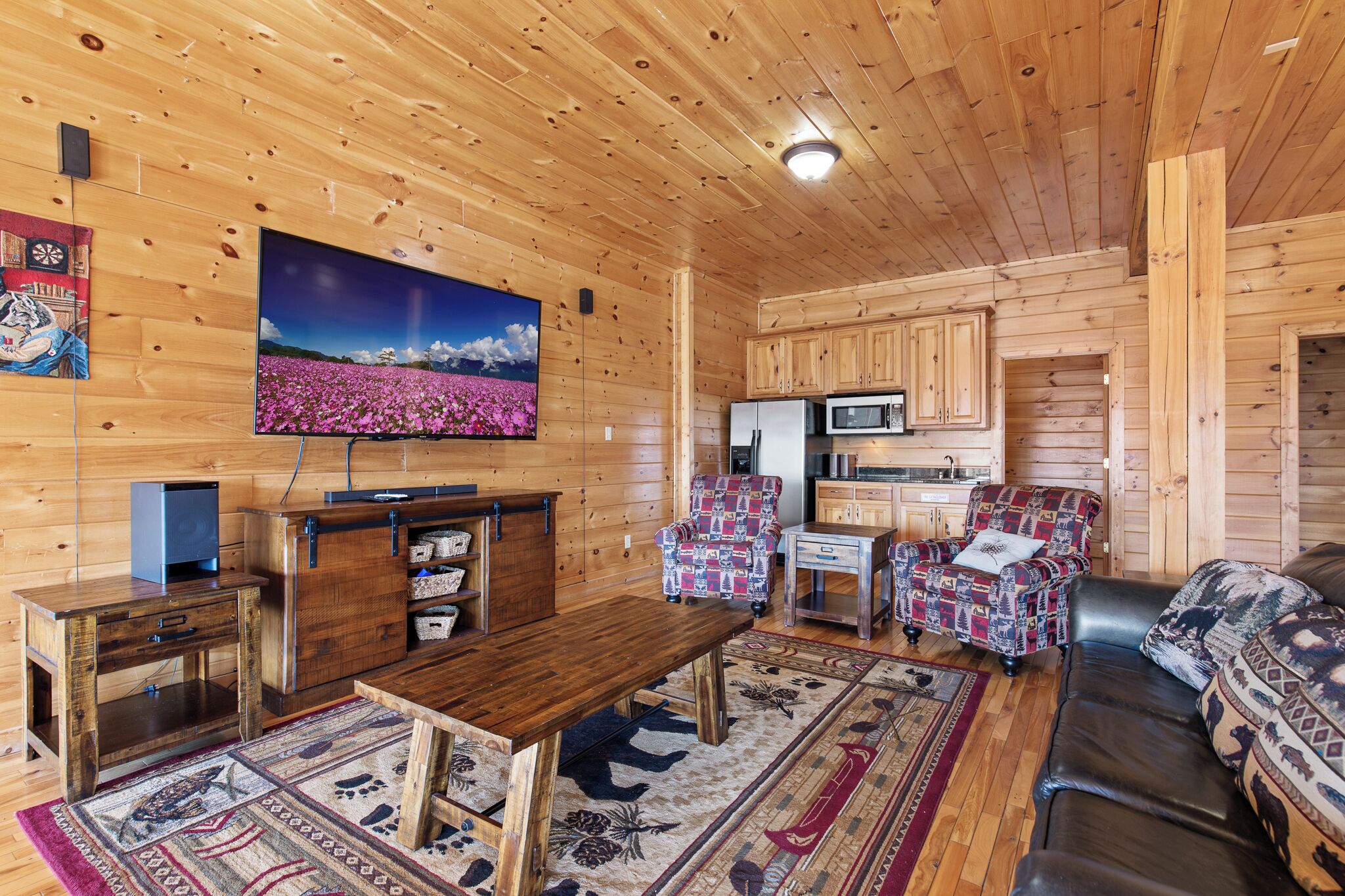 A cozy log cabin interior with a wooden dining table, a sofa, a patterned armchair, a flat-screen TV, and a kitchen area.