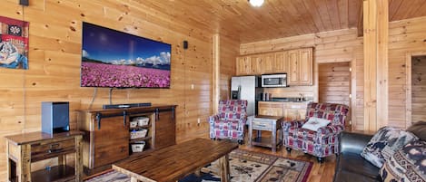 A cozy log cabin interior with a wooden dining table, a sofa, a patterned armchair, a flat-screen TV, and a kitchen area.
