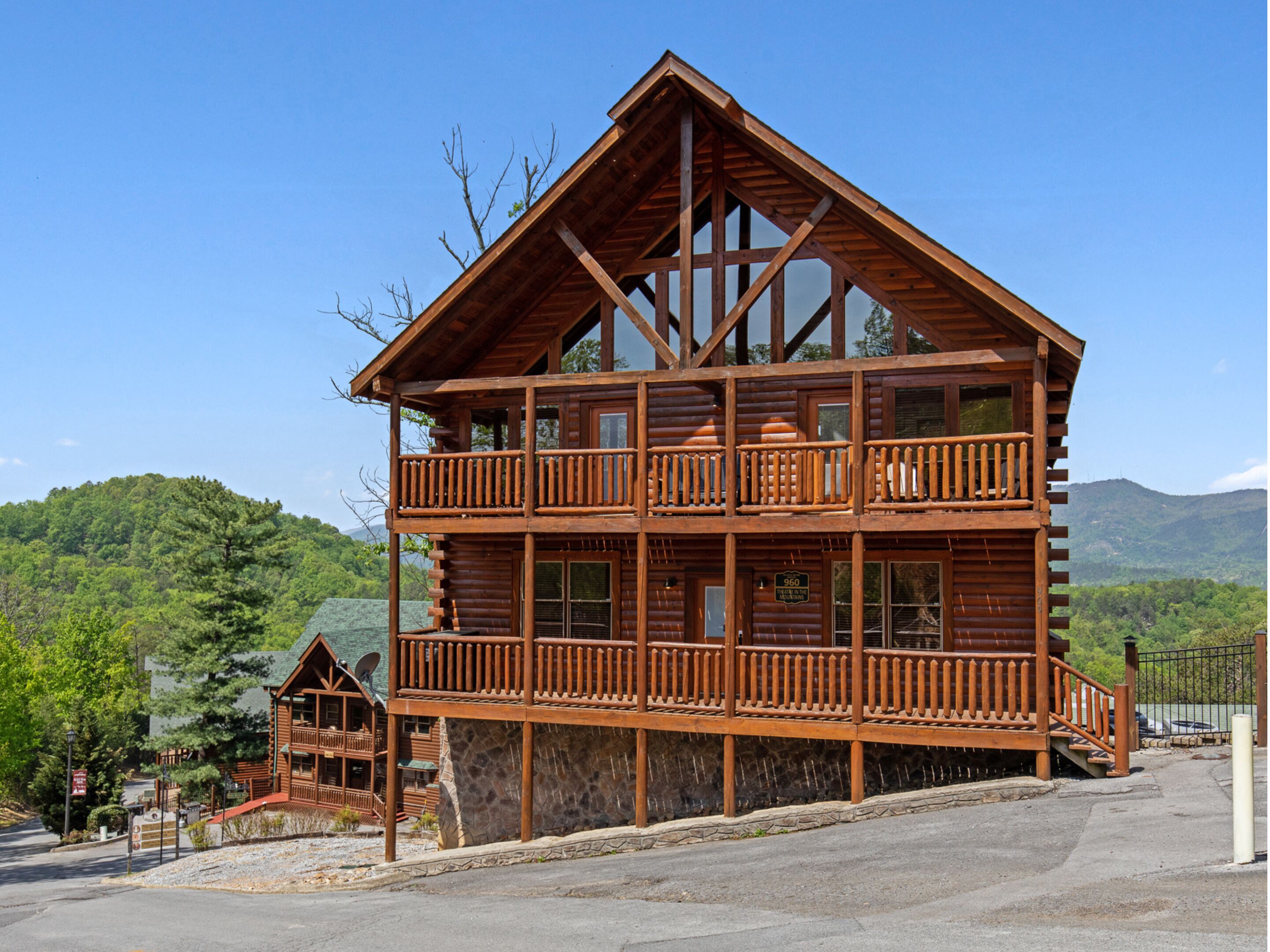 A two-story log cabin with a balcony, stone foundation, and a scenic mountain backdrop.