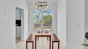 Un coin repas avec une table dressée pour deux personnes, un lustre moderne et une vue dégagée sur l’extérieur grâce à une grande fenêtre.