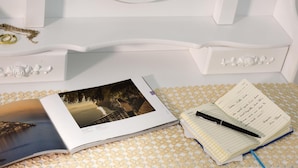 Una cama con cabecera blanca, un libro, una libreta y una pluma sobre un mantel estampado.