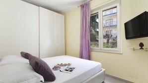 Un letto rifatto con cuscini, una finestra con tenda, una TV a parete e un piccolo ripiano con un oggetto decorativo.