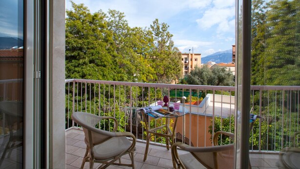 Un balcone con sedie in vimini, un tavolino con una rivista e vista su alberi e palazzi.