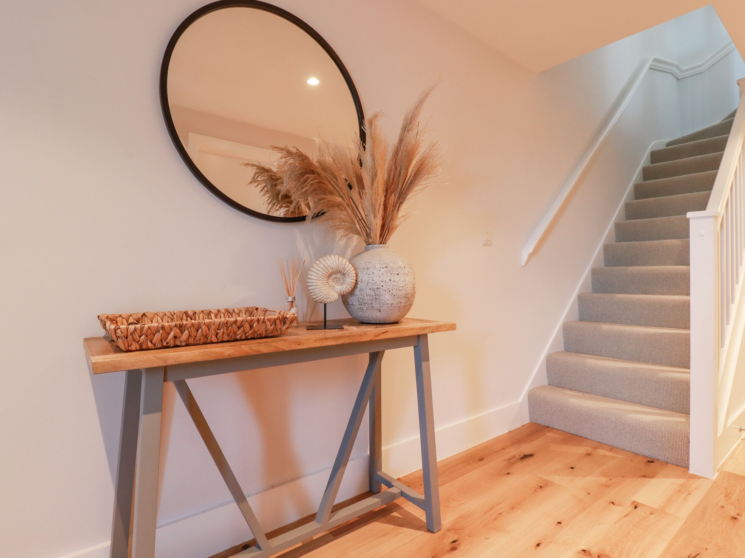 A wooden console table with a woven basket, a vase with pampas grass, and a decorative item. A round mirror hangs above. A staircase with carpeted steps is visible to the right.