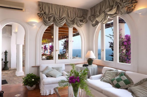 A cozy room with white furniture, large windows with curtains, and a view of the sea.