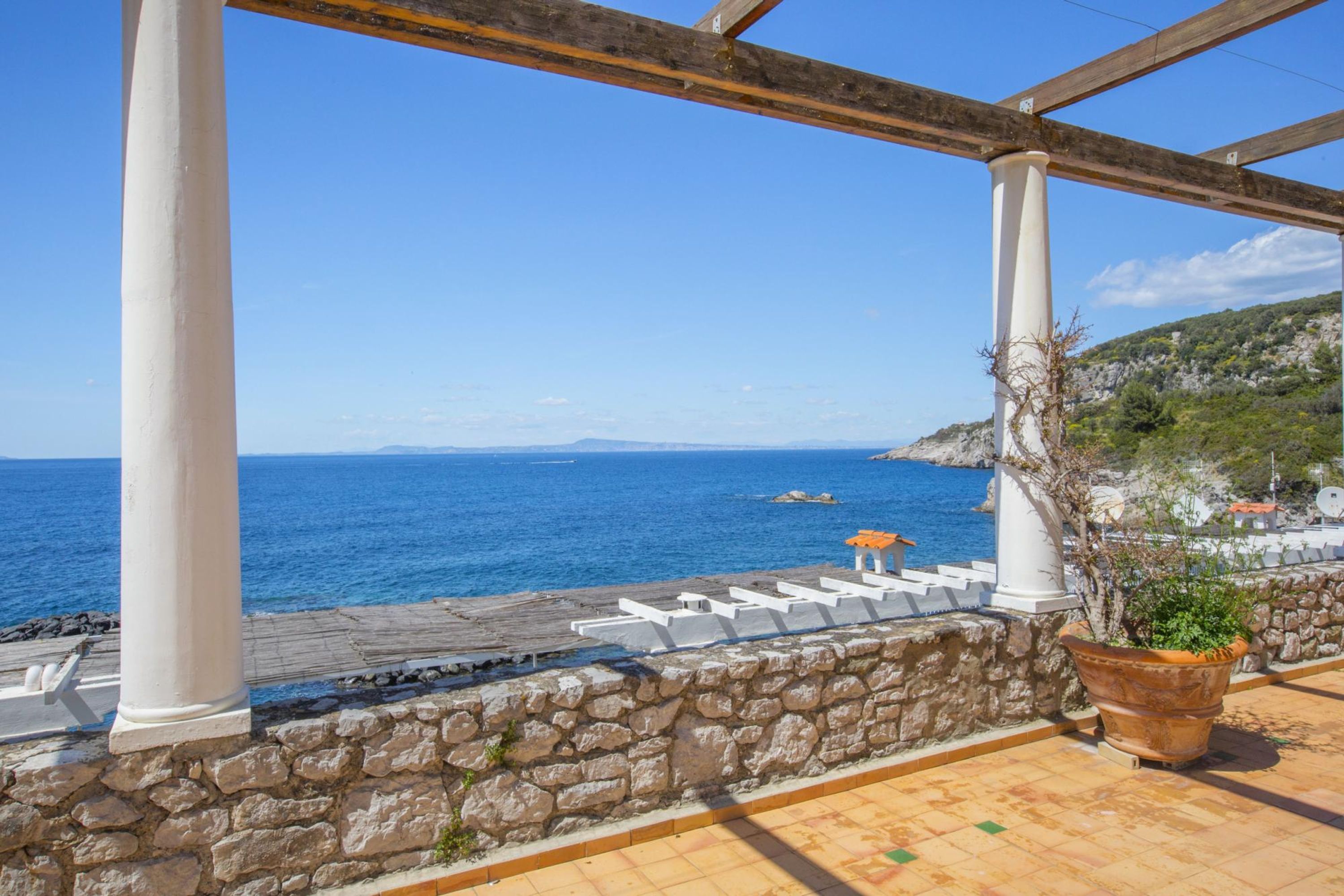 Eine Terrassenanlage am Meer mit Steinmauern, weißen Säulen und einem Blumentopf.