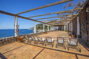 A covered outdoor seating area with chairs and potted plants, overlooking the sea.