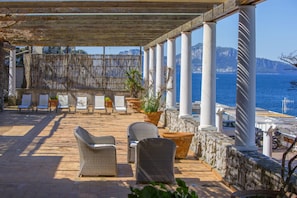 An outdoor patio with wicker furniture and potted plants, overlooking a coastal area with a stone wall and white columns.