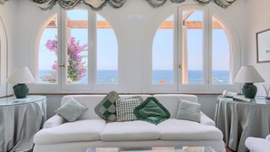 A white sofa with multiple cushions, a glass coffee table, and two lamps on side tables. Large arched windows offer a view of the sea. - Villa San Montano in Massa Lubrense (Massa Lubrense)