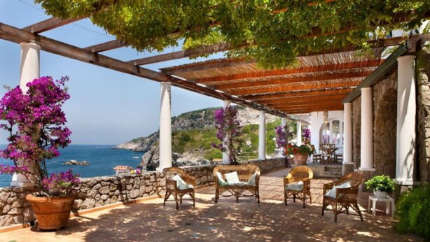 A covered outdoor seating area with wicker furniture, potted plants, and a view of the sea. - Villa San Montano in Massa Lubrense (Massa Lubrense)