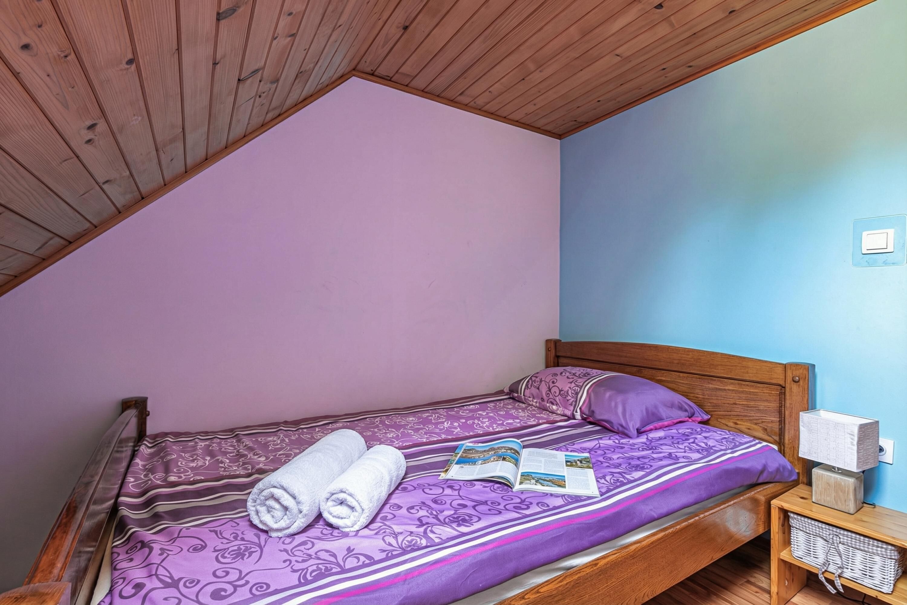 A single bed with a purple patterned bedspread, two rolled towels, and an open book on the bed.