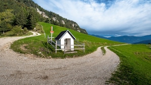 Een klein wit gebouw met een rood bord, omringd door een grindpad en groene velden, met bergen op de achtergrond.