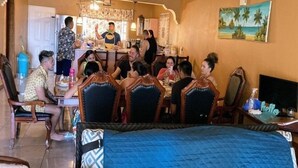 A dining area with wooden chairs and tables, a kitchen in the background, and a painting of a beach scene on the wall. - Yanceylargo Estate - Newly Renovated 7br Beautiful Two-story Estate Villa W Gorgeous Mountain View 7 Bedroom Villa by RedAwning (Montego Bay)