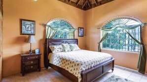 A bedroom with a wooden bed, a nightstand, and a window with a view of greenery. - Yanceylargo Estate - Newly Renovated 7br Beautiful Two-story Estate Villa W Gorgeous Mountain View 7 Bedroom Villa by RedAwning (Montego Bay)