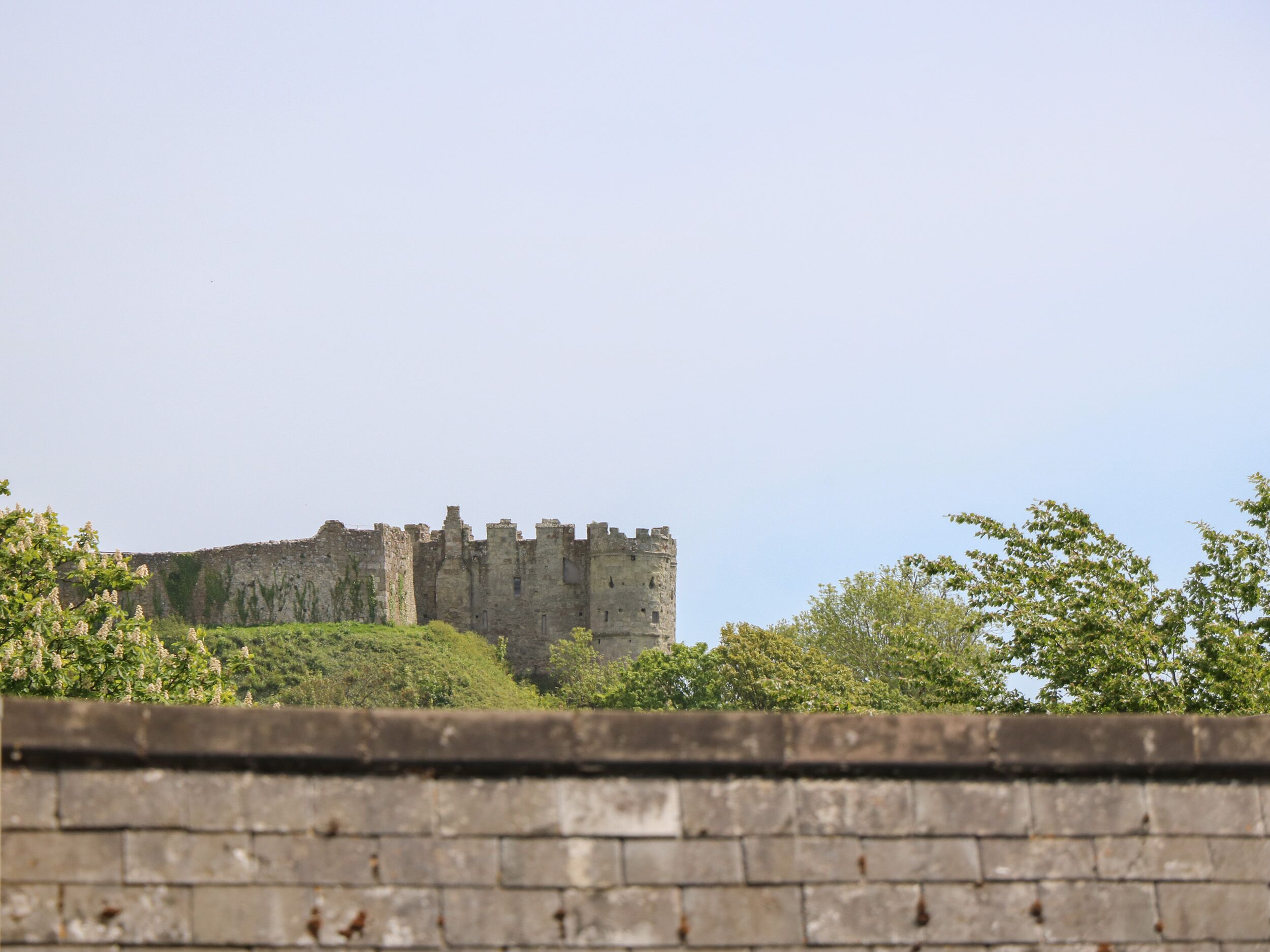 A historic stone castle with multiple towers and crenellations, surrounded by greenery and a low stone wall in the foreground.