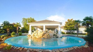 A swimming pool with a covered pavilion, surrounded by a landscaped garden with rocks and palm trees. - 2br/2ba Full Condo w/ Pool & Gym, Sleeps 6 (Gulf Shores)